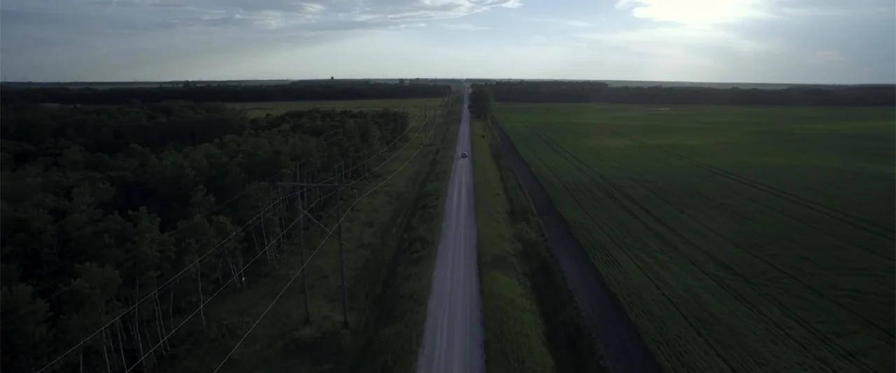 Radius (2017) movie still: establishing — A long straight asphalt road cuts through a vast rural landscape of fields. A small car travels down the…