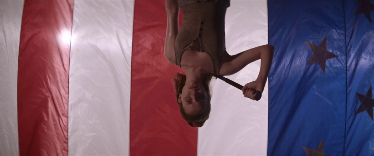 Assassination Nation (2018) movie still: medium — A woman is positioned upside down in front of a large American flag. She holds a baseball bat…