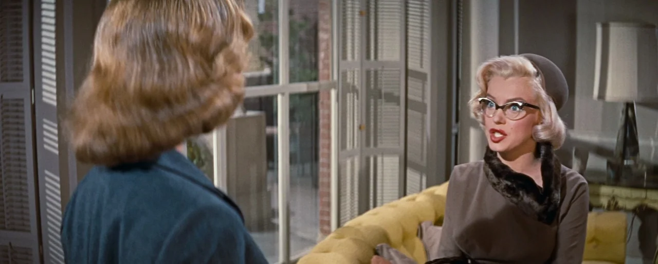 How to Marry a Millionaire movie still: medium — A 1950s interior space features a mustard yellow tufted sofa and a large window. One woman sits on…