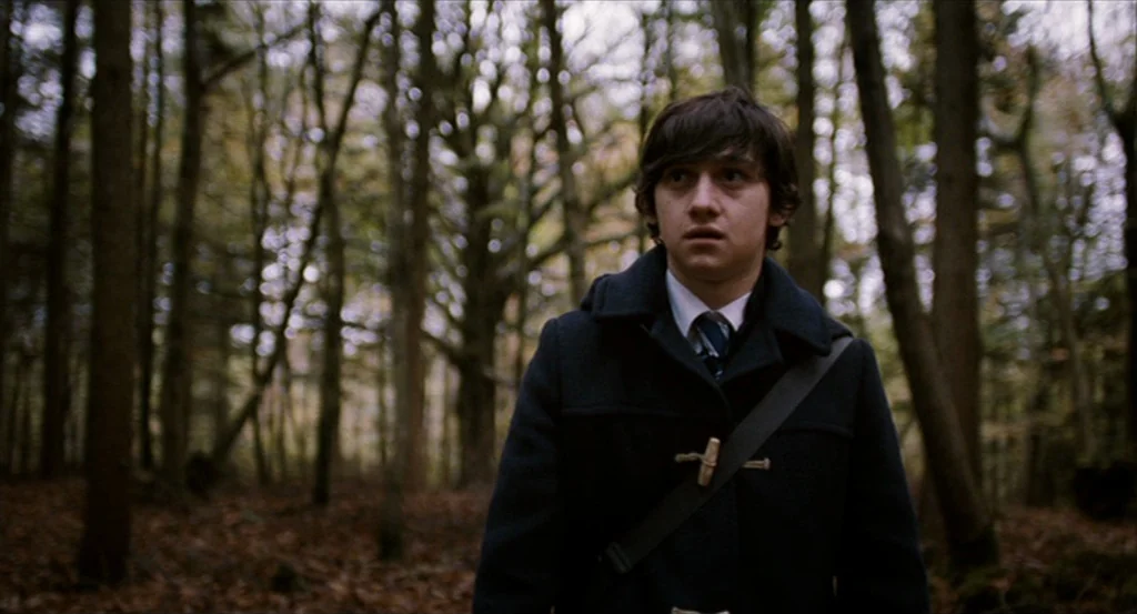 Submarine movie still: medium — A dense woodland with tall trunks and a ground covered in brown fallen leaves. A young man wearing a school uniform…