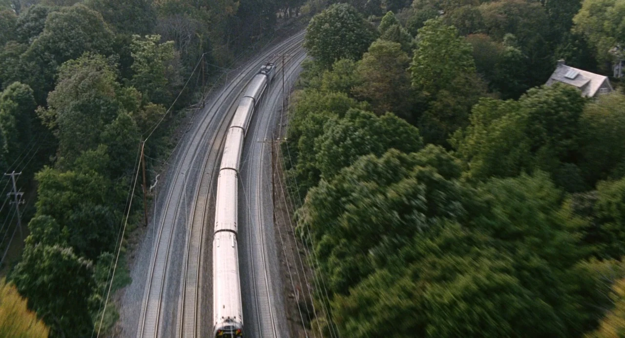 The Happening (2008) movie still: wide — A passenger train moves along tracks cutting through a dense green forest corridor. A single house with a…