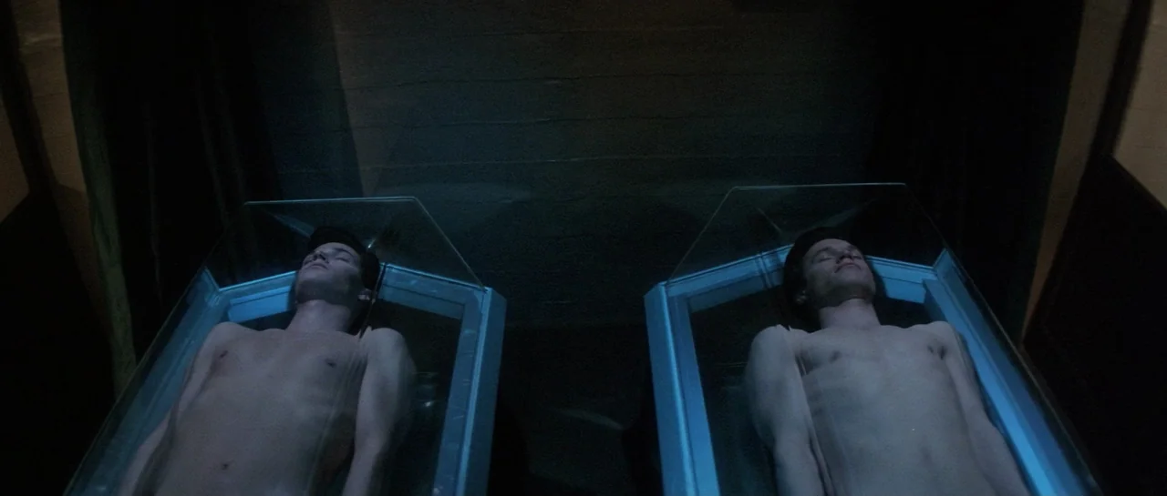 Lifeforce (1985) movie still: medium — A dark, spartan room contains two identical glass containers positioned side by side. Two young men lie…