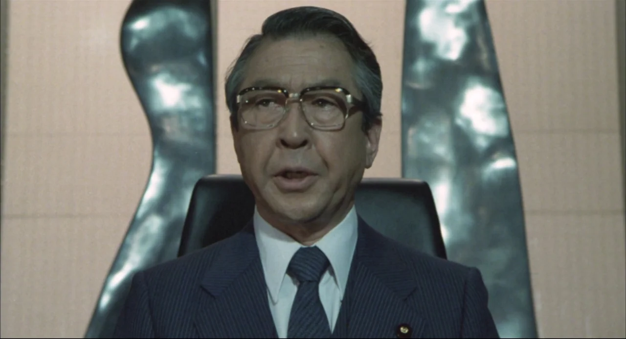 The Return of Godzilla (1984) movie still: medium — A man sits in a high-backed black chair in front of metallic decorative panels. He is wearing a…