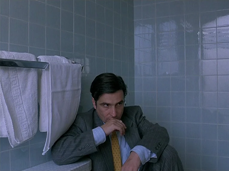 Detective (1985) movie still: medium — A man in a grey suit sits in a room with light blue square tiles. He rests his chin on his hand while seated…