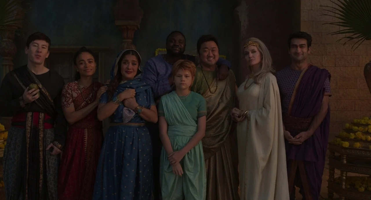 Eternals (2021) movie still: fullbody — A stone courtyard with palm plants and floral arrangements in the background. Eight people of various…