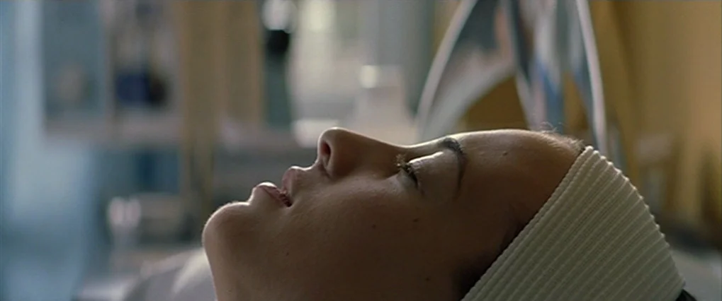 Talk To Her (2002) movie still: closeup — A clinical space with blurred, pale-toned medical equipment visible in the background. A woman in a white…