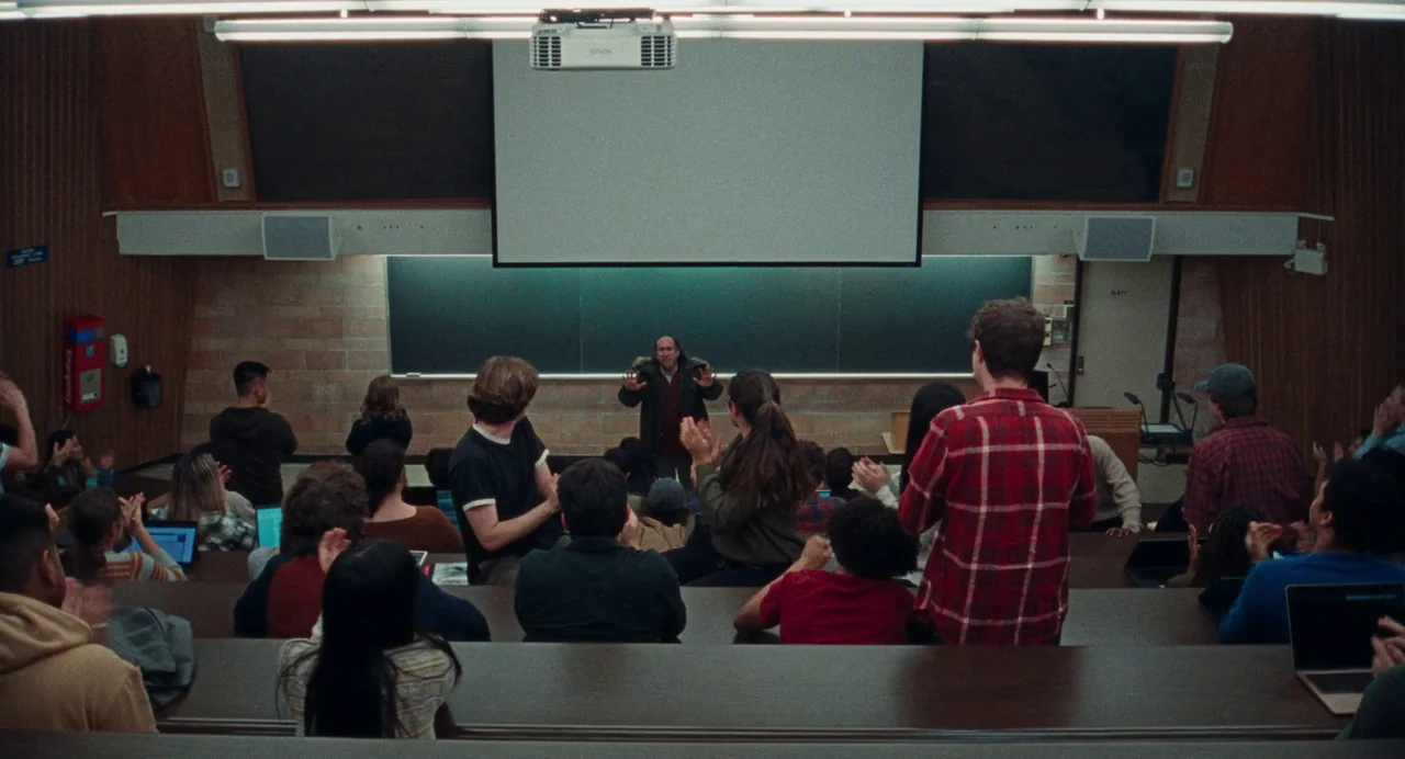 Dream Scenario (2023) movie still: wide — A tiered university lecture hall features a large black chalkboard and a projection screen. Students sit…