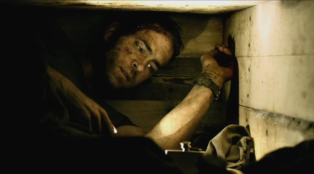 Buried (2010) movie still: closeup — A man lies trapped within the tight, wooden walls of a dark, narrow enclosure. He stares at a small metallic…