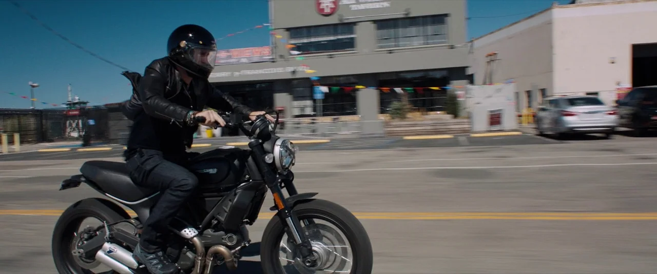 Venom (2018) movie still: fullbody — A man rides a motorcycle across an asphalt surface near a commercial building. Decorative bunting hangs from…