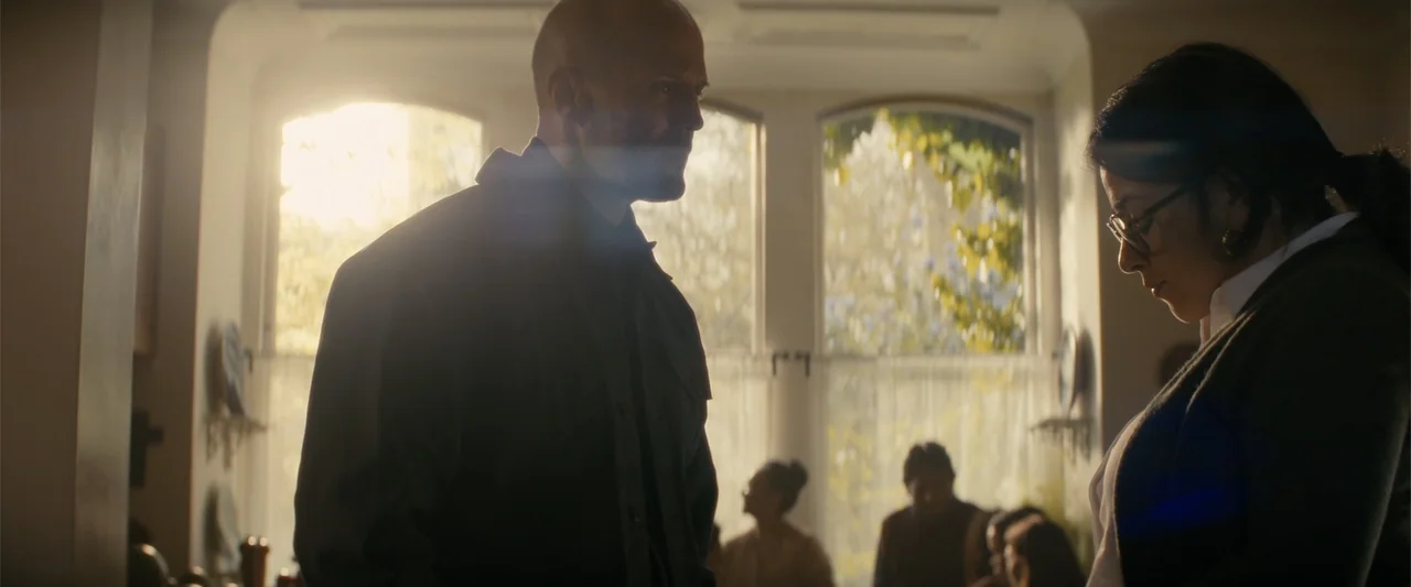 A Working Man (2025) movie still: medium — A well-lit interior living space with large windows and blurred figures in the distance. A man and woman…