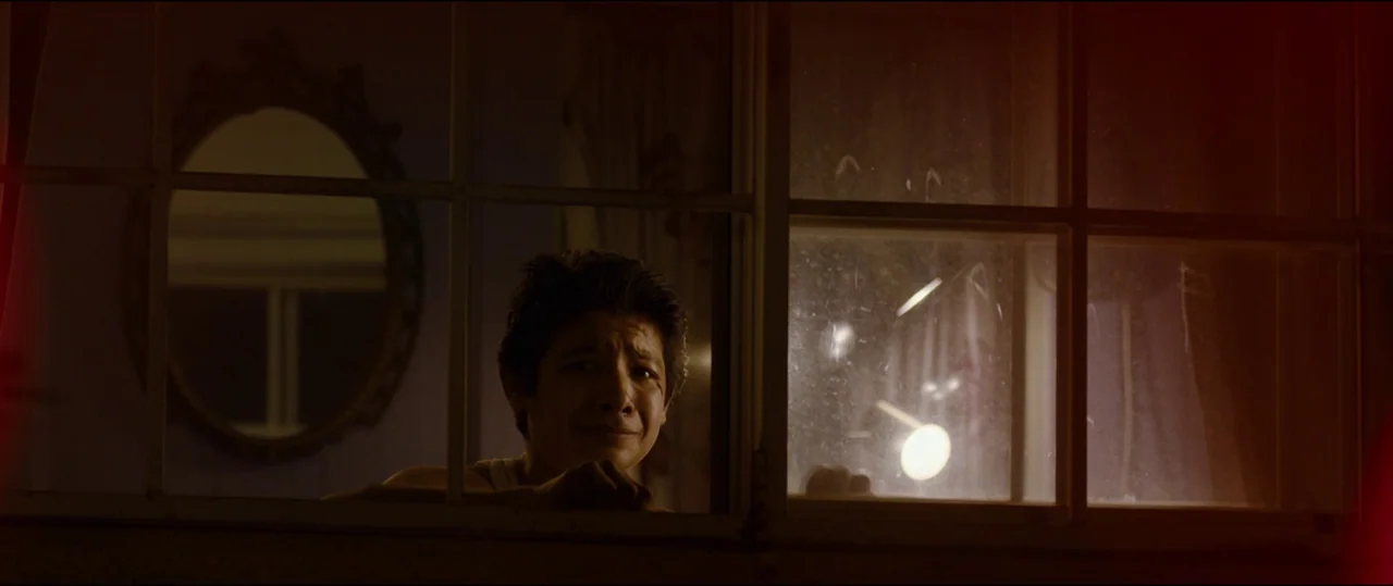 El Chicano (2018) movie still: medium — A person looks through a dirty window from an exterior, dark vantage point at night. The subject's face is…
