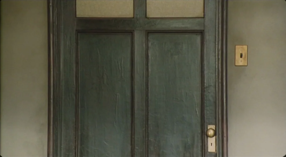 Madadayo (1993) movie still: medium — A dark wooden door with rectangular panels is centered within a plain grey wall. A brass doorknob and a…