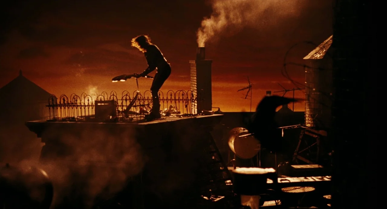 The Crow (1994) movie still: wide — An urban building top at night with a smoking chimney and water towers. A lone figure stands in silhouette on…