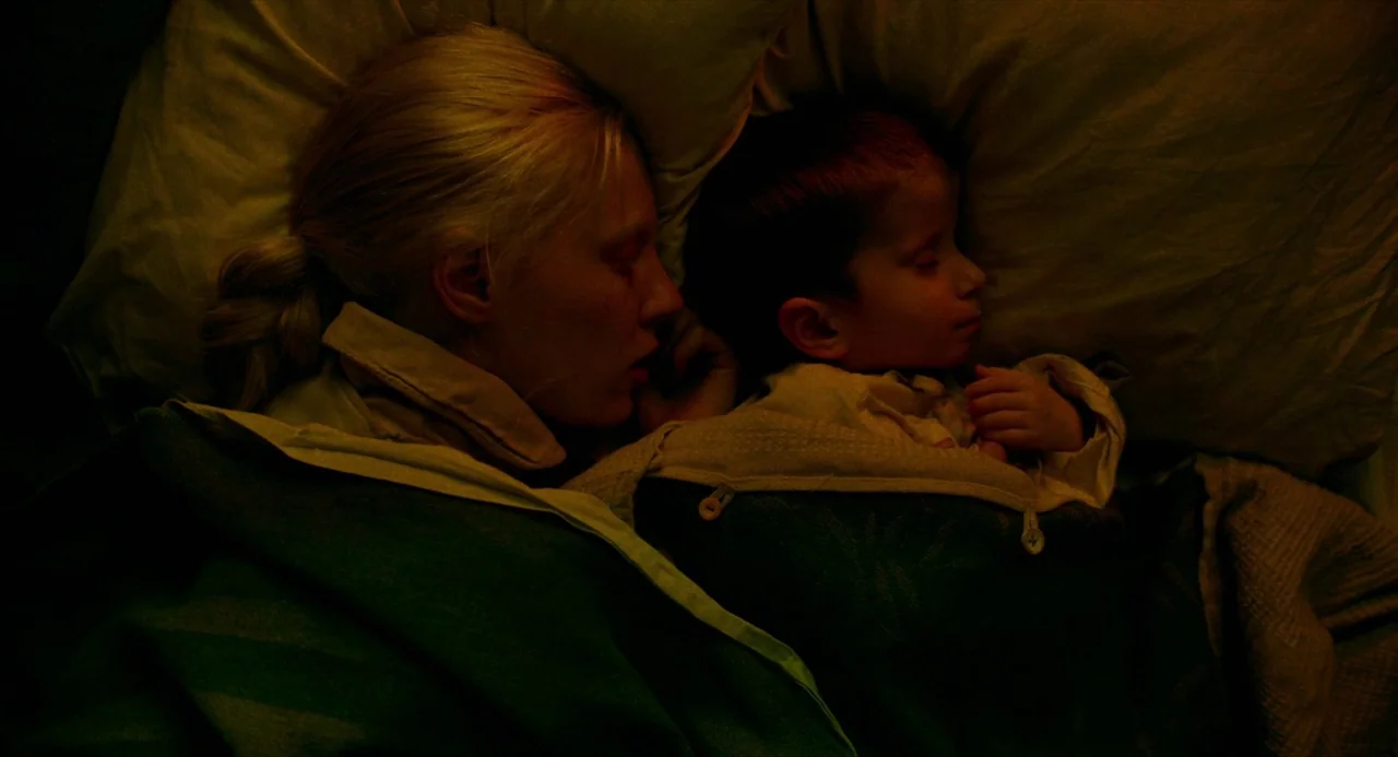 Beanpole (2019) movie still: closeup — A woman and a young child sleep in a dark room under blankets. They lie closely together in bed with their…