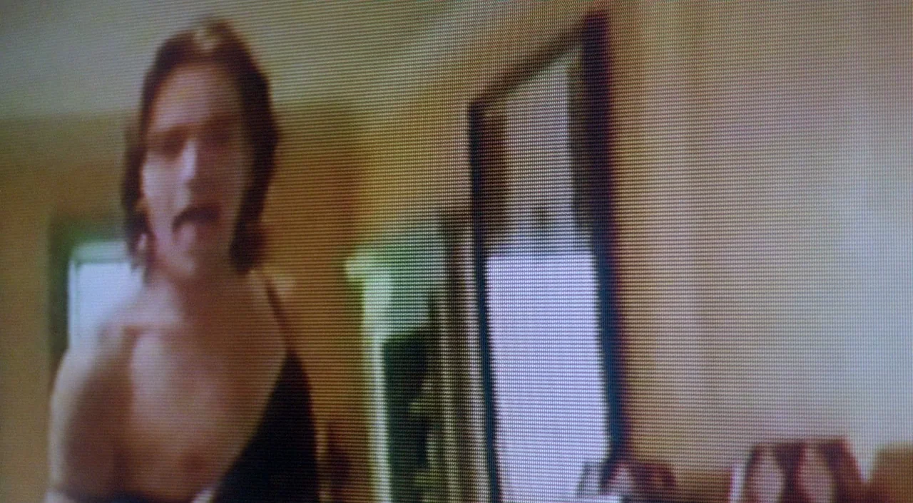 Shallow Grave (1994) movie still: medium — A dimly lit indoor space with blurred furniture and a mirror frame in back. A man with shoulder-length…