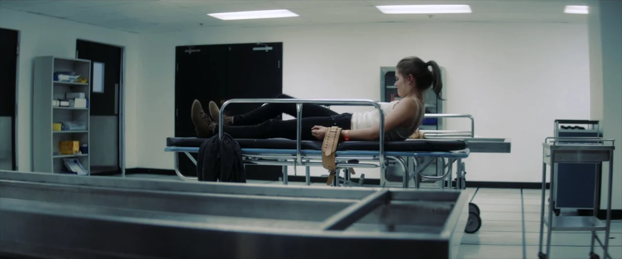 Tell Me How I Die (2016) movie still: medium — A sterile morgue with white walls, tiled floors, and stainless steel autopsy tables. A woman in a…