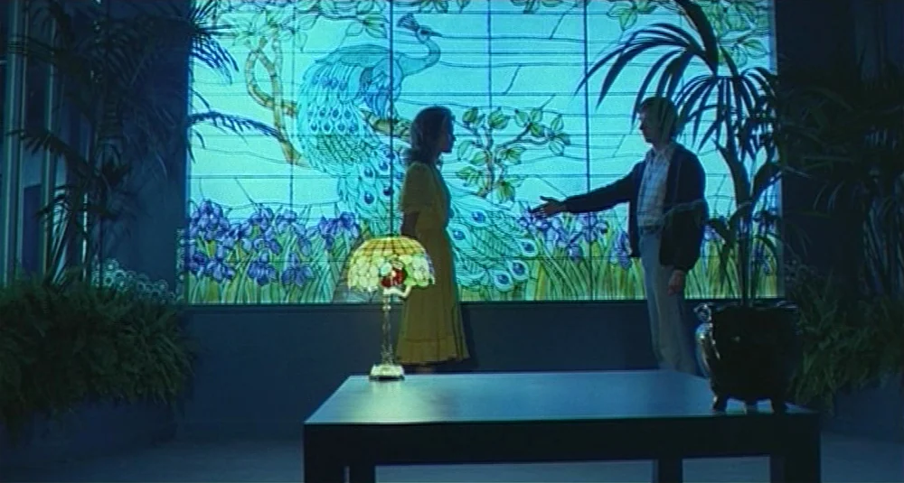 Footprints (Le Orme) (1975) movie still: fullbody — A spacious room features a large, backlit stained glass display depicting a peacock. A man and a…
