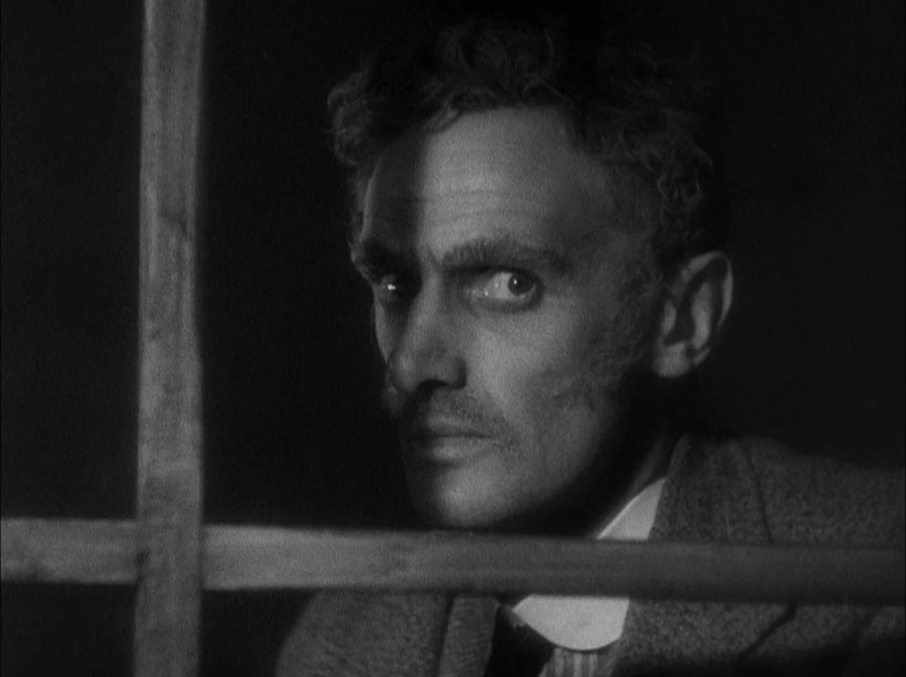The 39 Steps (1935) movie still: closeup — A dark, cramped space with wooden bars foregrounding the subject. The man looks toward the camera with…