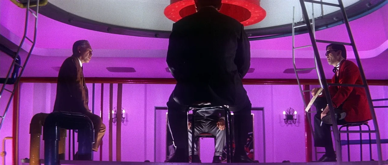 Tokyo Drifter (1966) movie still: fullbody — A spacious, sterile room features bright purple walls and metal industrial piping structures. Three men…