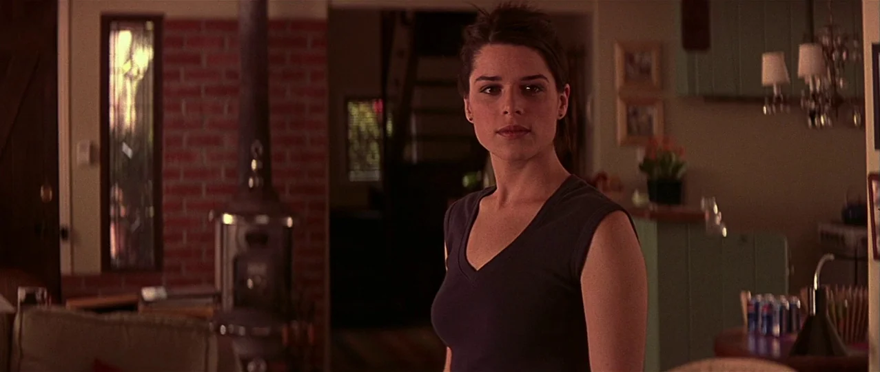 Scream 3 (2000) movie still: medium — An open plan residential interior features a red brick wall and a wood-burning stove. A young woman stands in…