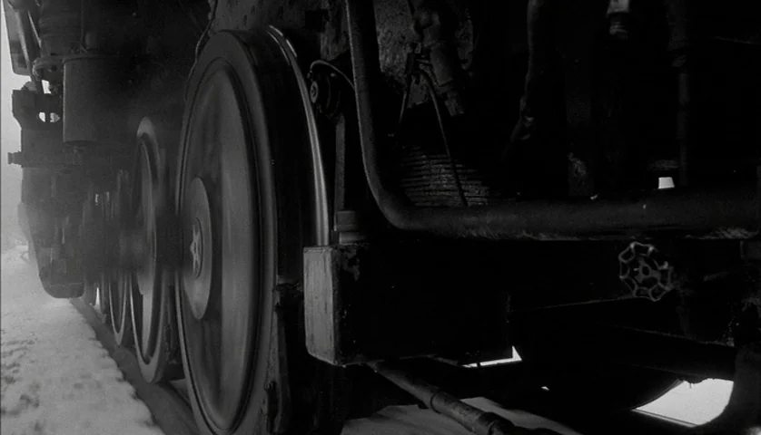 Dead Man (1995) movie still: closeup — The rotating steel wheels of a massive steam locomotive move against a snowy ground. Mechanical levers and…
