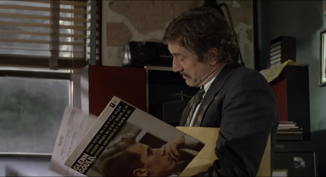 Cop Land (1997) movie still: medium — An office interior features a window with horizontal blinds and a map on the wall. A man in a suit looks down…