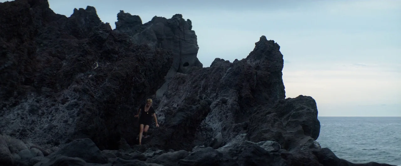 Sibyl (2019) movie still: wide — A rugged volcanic coastline features massive, dark, porous rock formations meeting a calm ocean. A woman in a black…