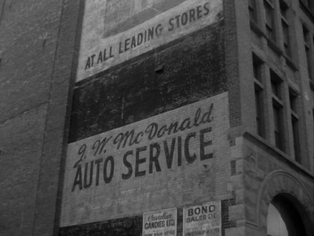My Winnipeg (2007) movie still: wide — The exterior side of a multi-story urban brick building features large painted advertisements. Large black…