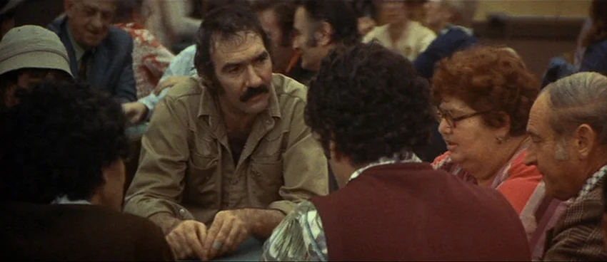 California Split (1974) movie still: medium — A crowded public dining space with multiple people sitting at tables in mid-conversation. A man in a…
