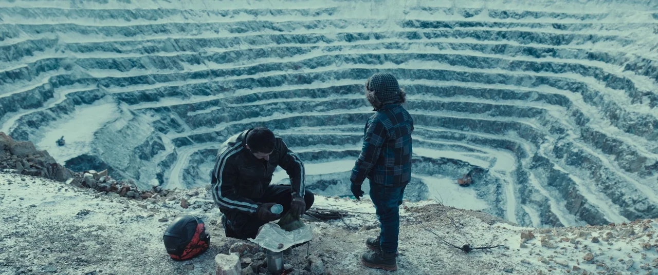 R.M.N. (2022) movie still: wide — A deep, snow-covered open pit mine stretches out behind two people. An adult man and a child stand on the edge of…