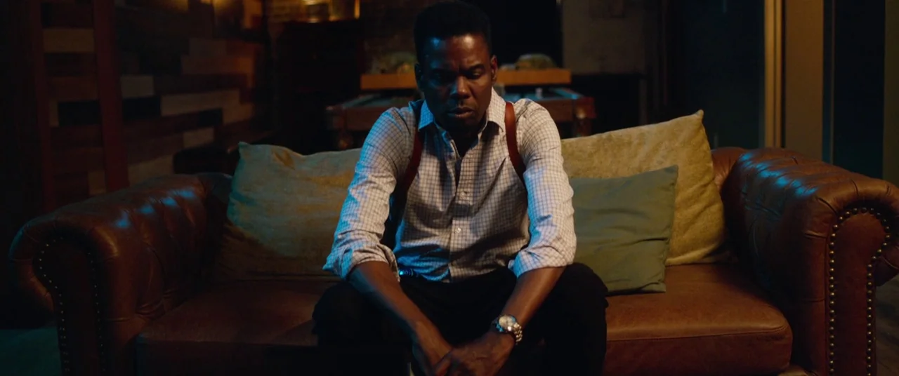 Spiral (2021) movie still: medium — A dimly lit room contains a large brown leather sofa with green cushions. A man in a light plaid shirt sits on…