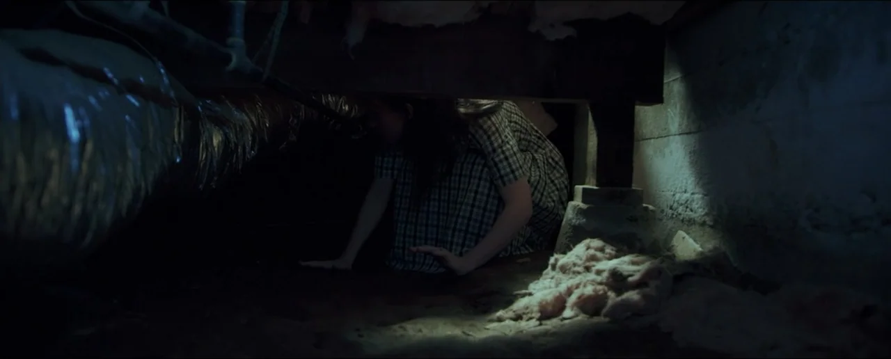 Hostages (2017) movie still: medium — A dark, cramped crawlspace filled with dirt, wooden beams, and insulation materials. A woman in a plaid dress…