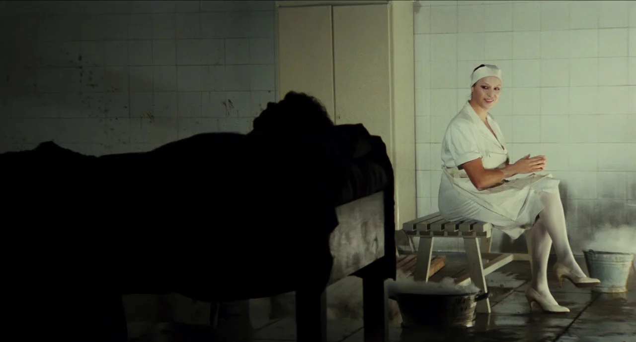 City of Women (1980) movie still: medium — A tiled room contains a metal cart holding a shrouded figure in deep shadow. A nurse in a white uniform…