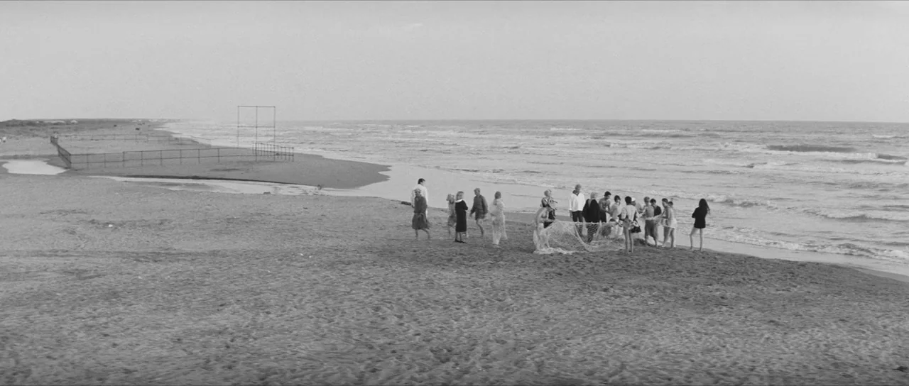 La Dolce Vita (1960) movie still: wide — A wide sandy beach stretches toward an overcast horizon with calm waves crashing. A group of people stands…