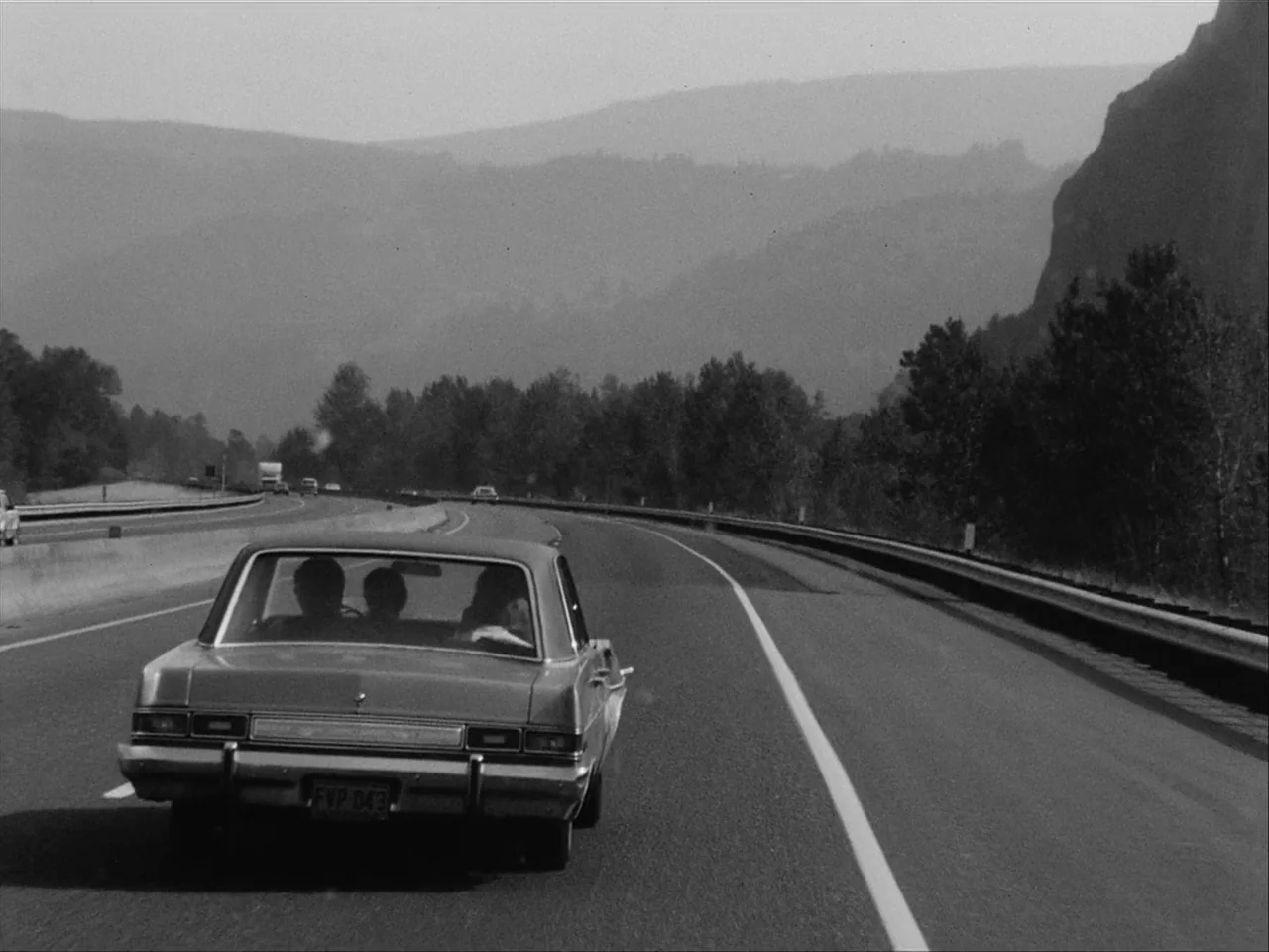 Mala Noche (1986) movie still: wide — A rural highway stretches forward beneath rolling mountains under a clear sky. A vintage car moves along the…