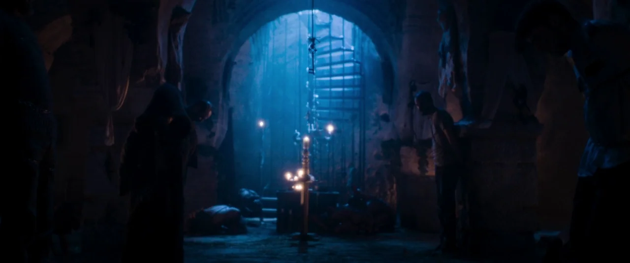 Baskin (2015) movie still: wide — A dark subterranean stone chamber features arched ceilings and a spiral staircase in back. Several figures stand…