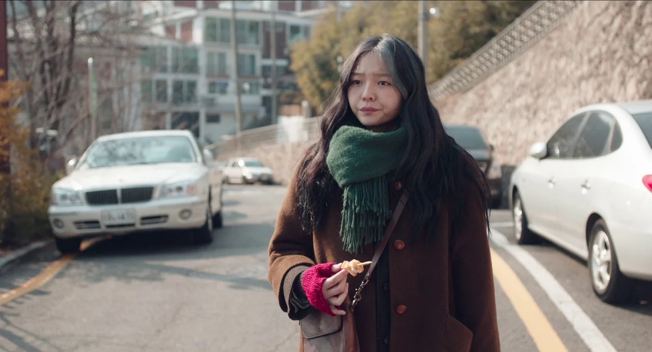 Microhabitat (2017) movie still: medium — An asphalt road in an urban area features parked white vehicles on both sides. A young woman walks through…