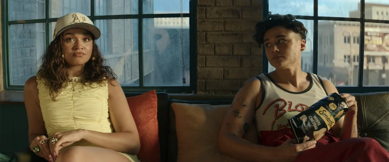 Maintenance Required (2025) movie still: medium — An interior space with a black leather sofa positioned against a dark grid window. Two women sit…