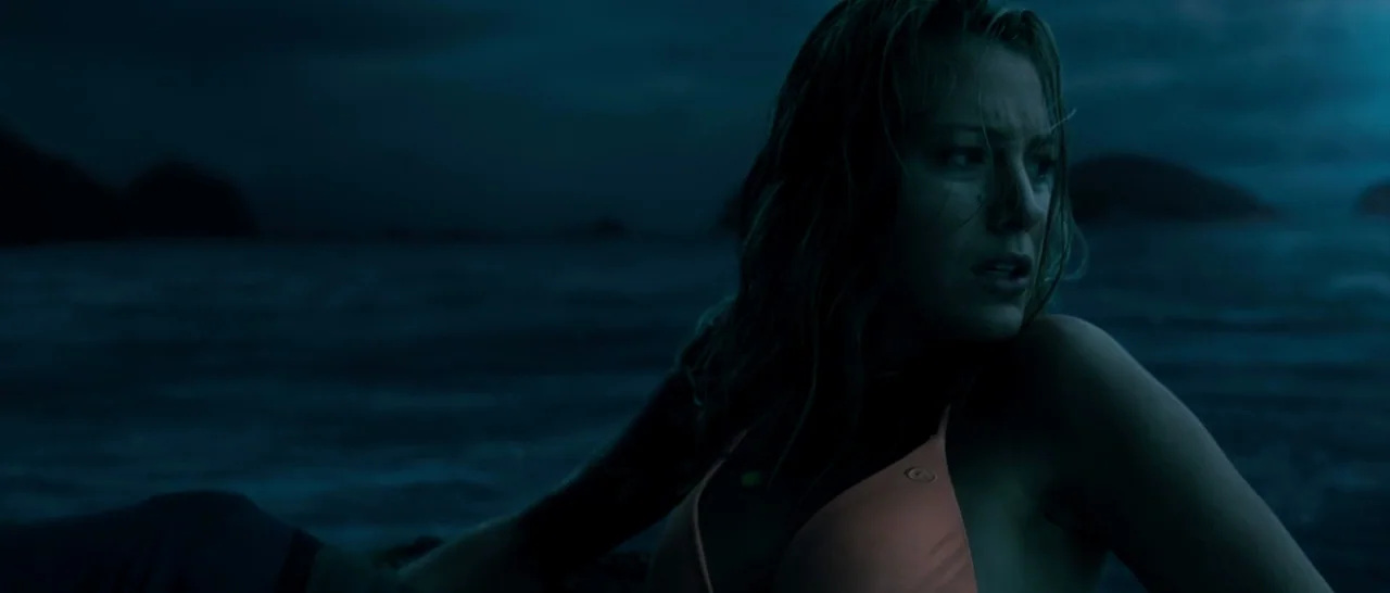 The Shallows (2016) movie still: medium — A dark rocky outcrop sits surrounded by turbulent water at night under a dim sky. A woman with wet blonde…