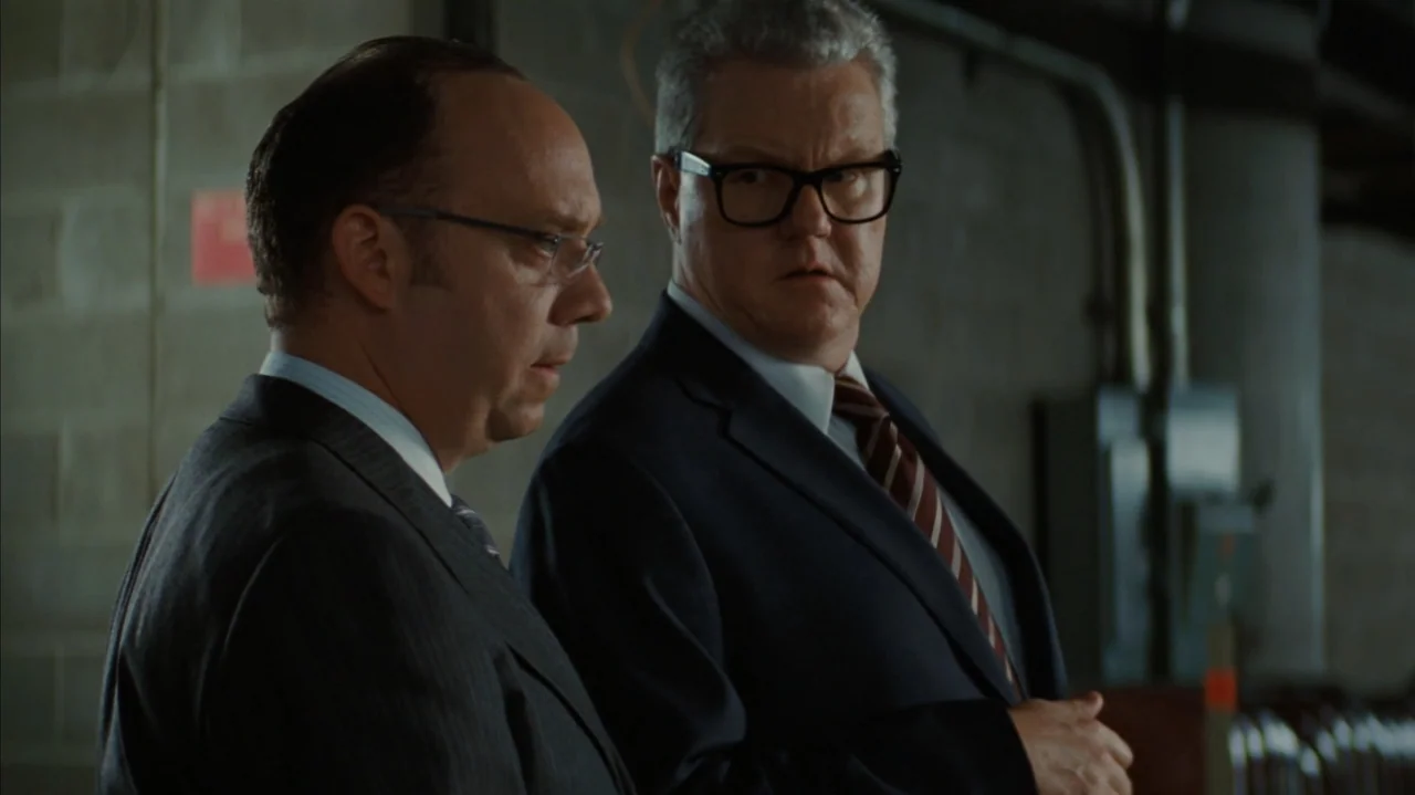 Duplicity (2009) movie still: medium — An interior industrial space featuring grey cinder block walls and metallic equipment components. Two men in…