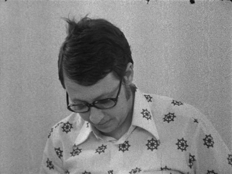 Family Nest (1979) movie still: closeup — A man in a patterned button-down shirt sits against a plain, light-colored background. He looks down…