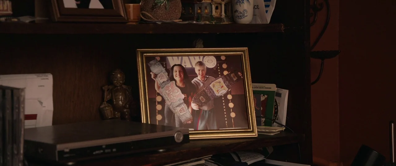 Fighting With My Family (2019) movie still: closeup — A wooden shelf holds a collection of domestic objects including a framed childhood photograph.…