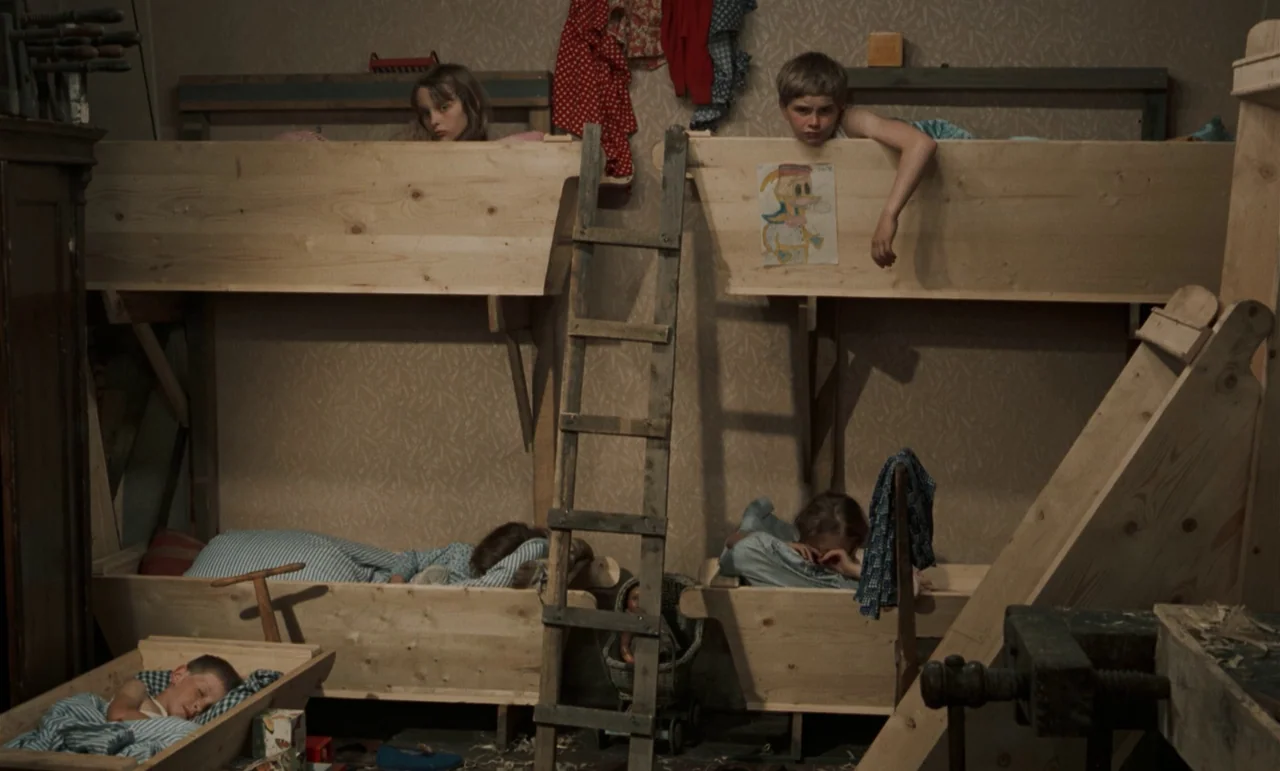 Larks On A String (1969) movie still: wide — A room contains multiple stacked wooden bunk beds and a central ladder. Four children lay in the beds…