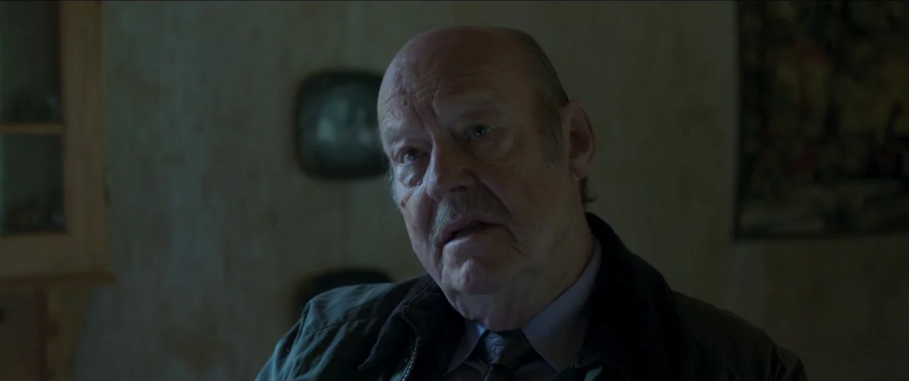 Border movie still: closeup — An interior space features muted wallpaper and minimal visible decor in deep shadows. An elderly man with a mustache…