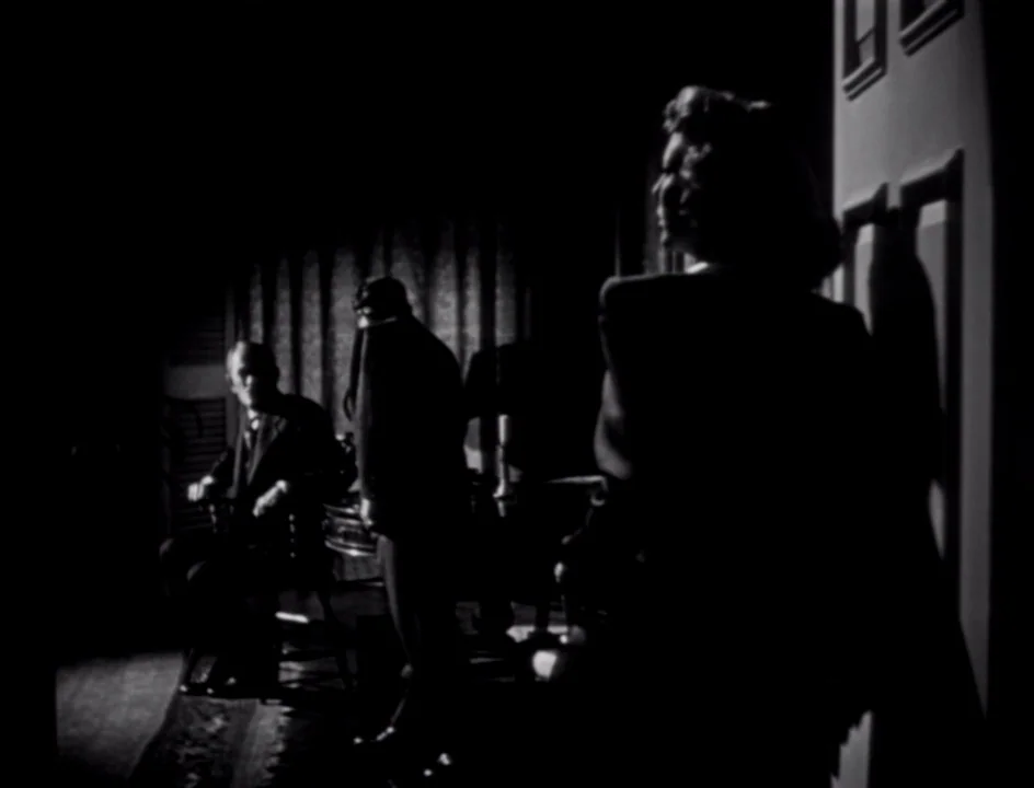 The Stranger (1946) movie still: medium — A dark study with heavy curtains and a wooden chair in low key light. A woman is positioned in the…