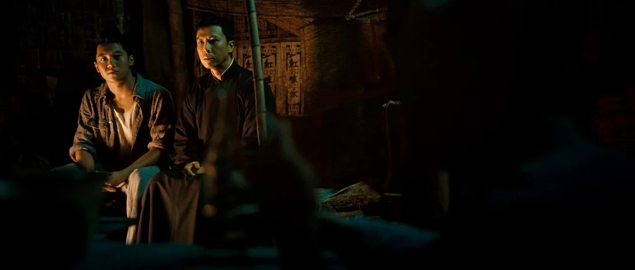 Ip Man 2 (2010) movie still: medium — A rustic interior with woven reed walls and dim, localized warm lighting. Two men sit side by side in the…