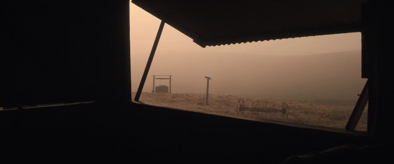 The New Boy (2023) movie still: establishing — A dark interior structure frames an open field filled with hazy golden light. An old metal well and a…