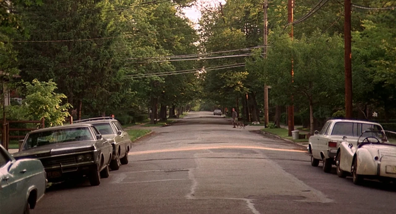 Taking Off (1971) movie still: establishing — A suburban residential street is lined with mature trees and parked vintage automobiles. A person…