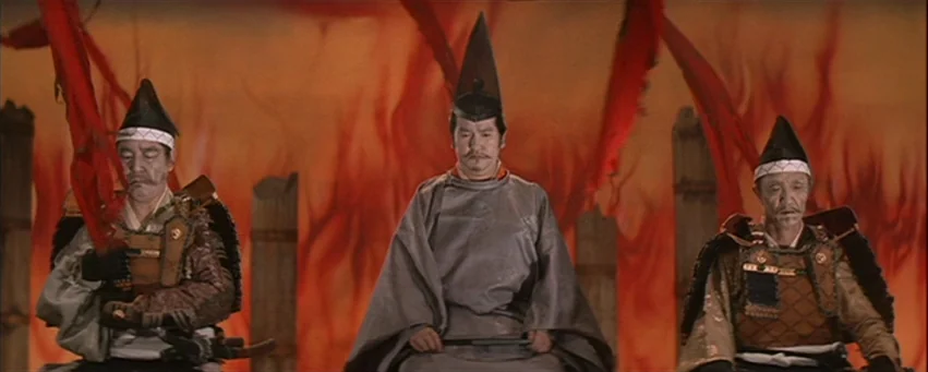 Kwaidan (1964) movie still: medium — Three Japanese men sit in a row before a backdrop of painted flames. The central figure wears a plain gray…