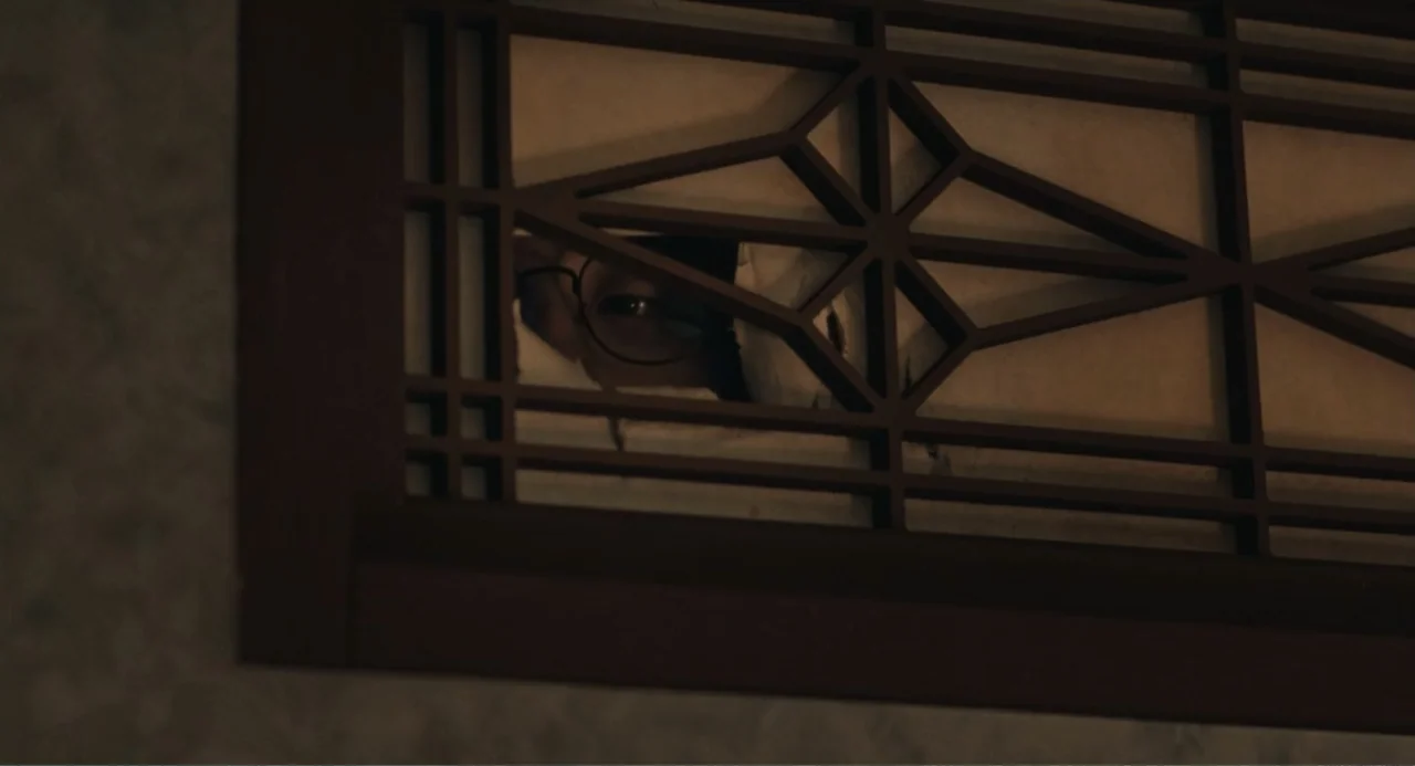 Winter in Sokcho (2025) movie still: closeup — A traditional wooden lattice screen with paper inserts is partially torn in the center. An eye…